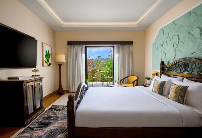 King bedroom with ornate wooden bed and balcony garden views at Mercure Goa Devaaya Resort